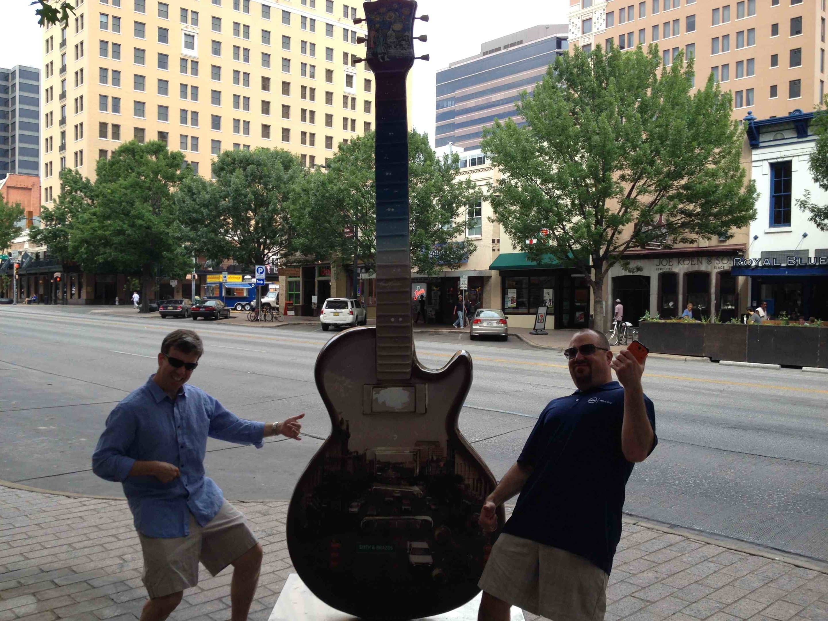 Two men doing air guitar next to giant guitar.