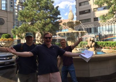Three team members stand in front of a fountain, smiling as they raise their hands during a team building activity in Toronto Bloor Yorkville.