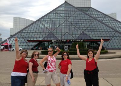 Five women standing in front of the Rolling Stones building, raising their hands and smiling during a team building activity in Cleveland.