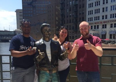 Three people are taking a photo while making a thumbs-up gesture alongside a sculpture doing the same thumbs-up pose