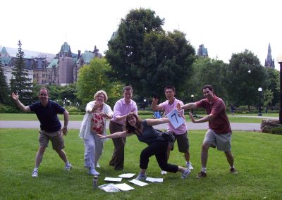 A team posing in a field, with five members at the back extending their hands forward toward a girl in the front, who is striking a full-body pose during a team building activity in Ottawa.