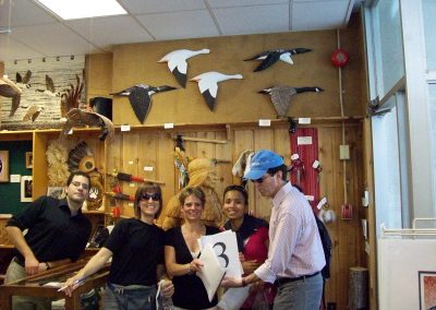 A scavenger hunt team of 5 are standing in an antique shop, surrounded by various art pieces, with a Native American sculpture featured in the background.