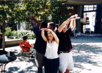 Five individuals are positioned in a circle, each standing with their backs pressed against one another while reaching upward with their hands raised during a team building activity in Seattle Downtown.